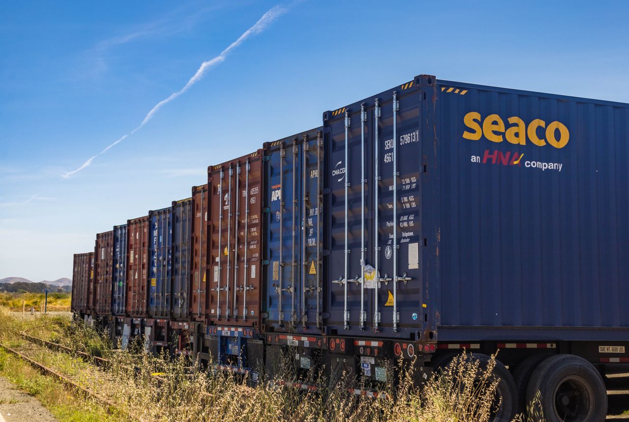Home Row of vibrant cargo containers on train tracks under a clear blue sky.
